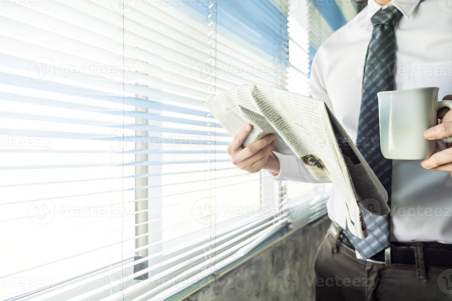 Businessman reading newspaper and holding coffee cup while standing at a window in an office. photo