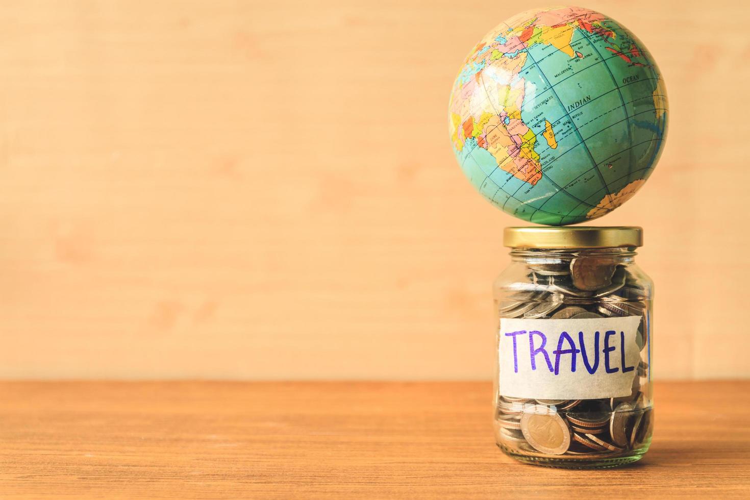 Coins in glass jar with travel label and globe on wooden table. Saving money for travel concept. Free space for text photo