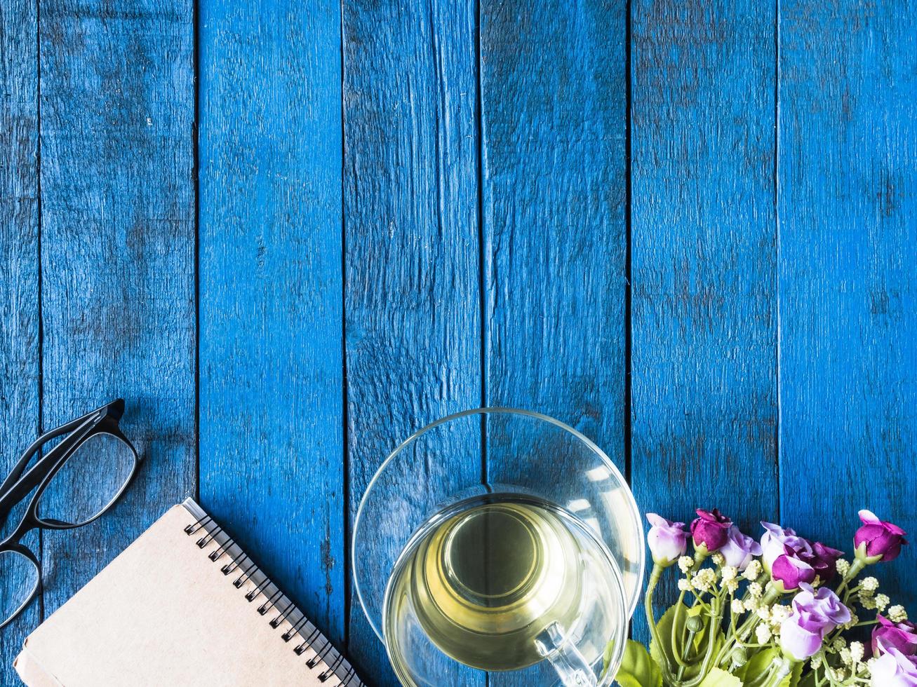 Top view of tea cup with flower, notebook and glasses on blue wooden table background. Free space for text photo