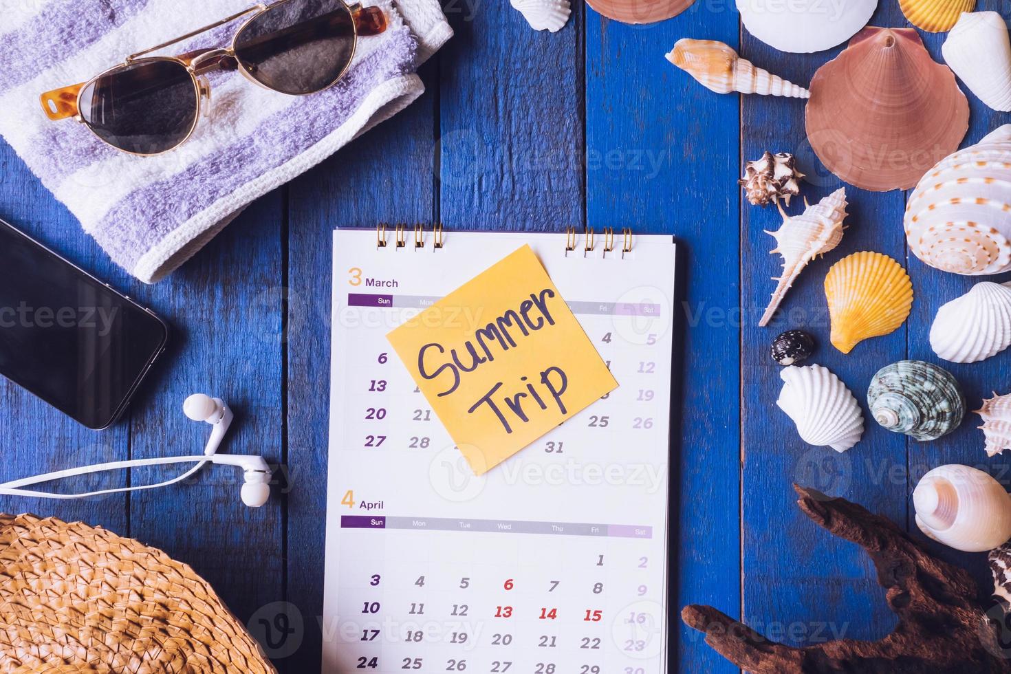 Top view of calendar with accessories for summer trip and seashells on blue painted wood plank background. photo