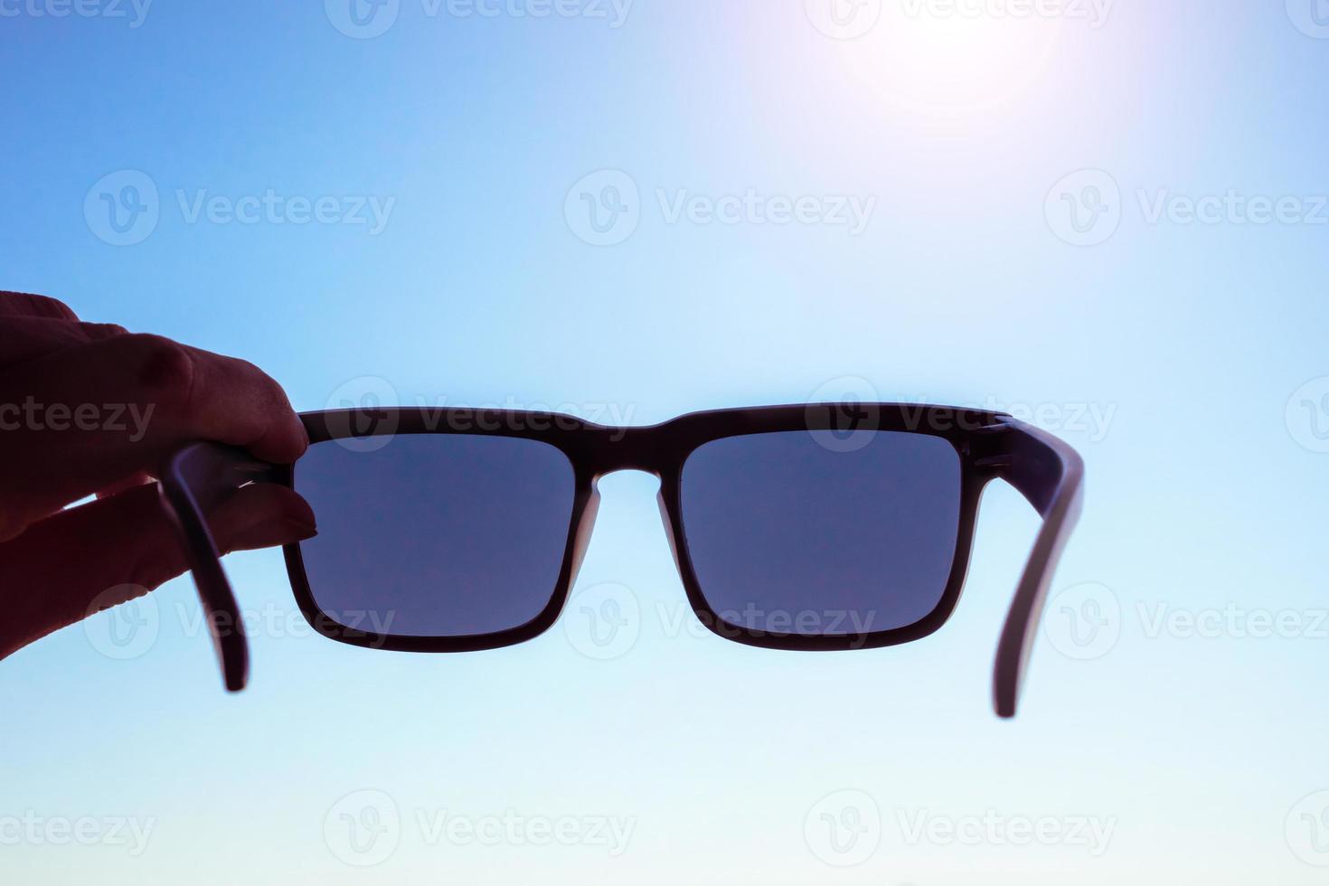 mano masculina sosteniendo gafas de sol frente al sol, cielo azul en el fondo foto