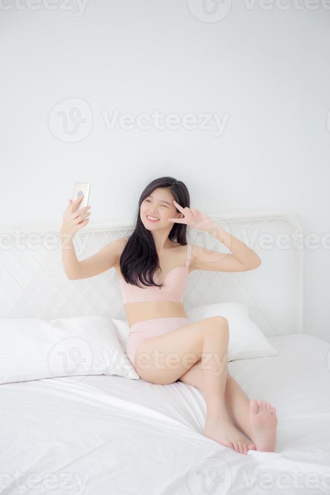 hermosa joven asiática sexy en ropa interior hablando un selfie en un teléfono inteligente para la red social en el dormitorio, una chica en lencería se relaja tomando una foto en el teléfono móvil en la cama en el dormitorio.