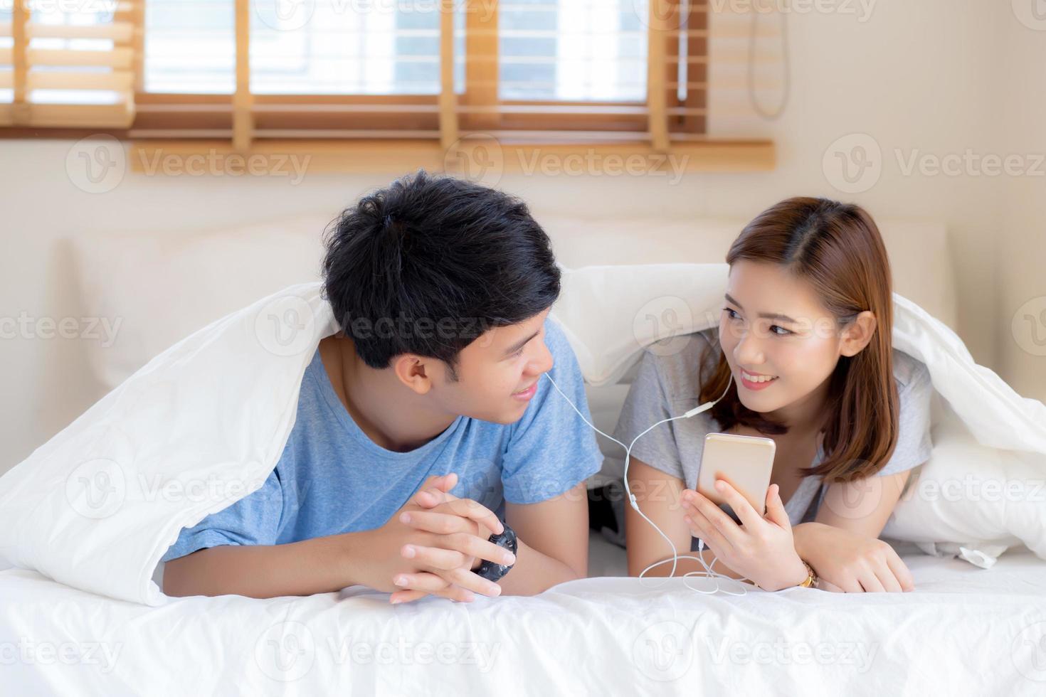 Young asian couple smile listen music with smart mobile phone on bed at bedroom with fun and enjoy, family sharing podcast or watching video from internet online with relax, lifestyle concept. photo