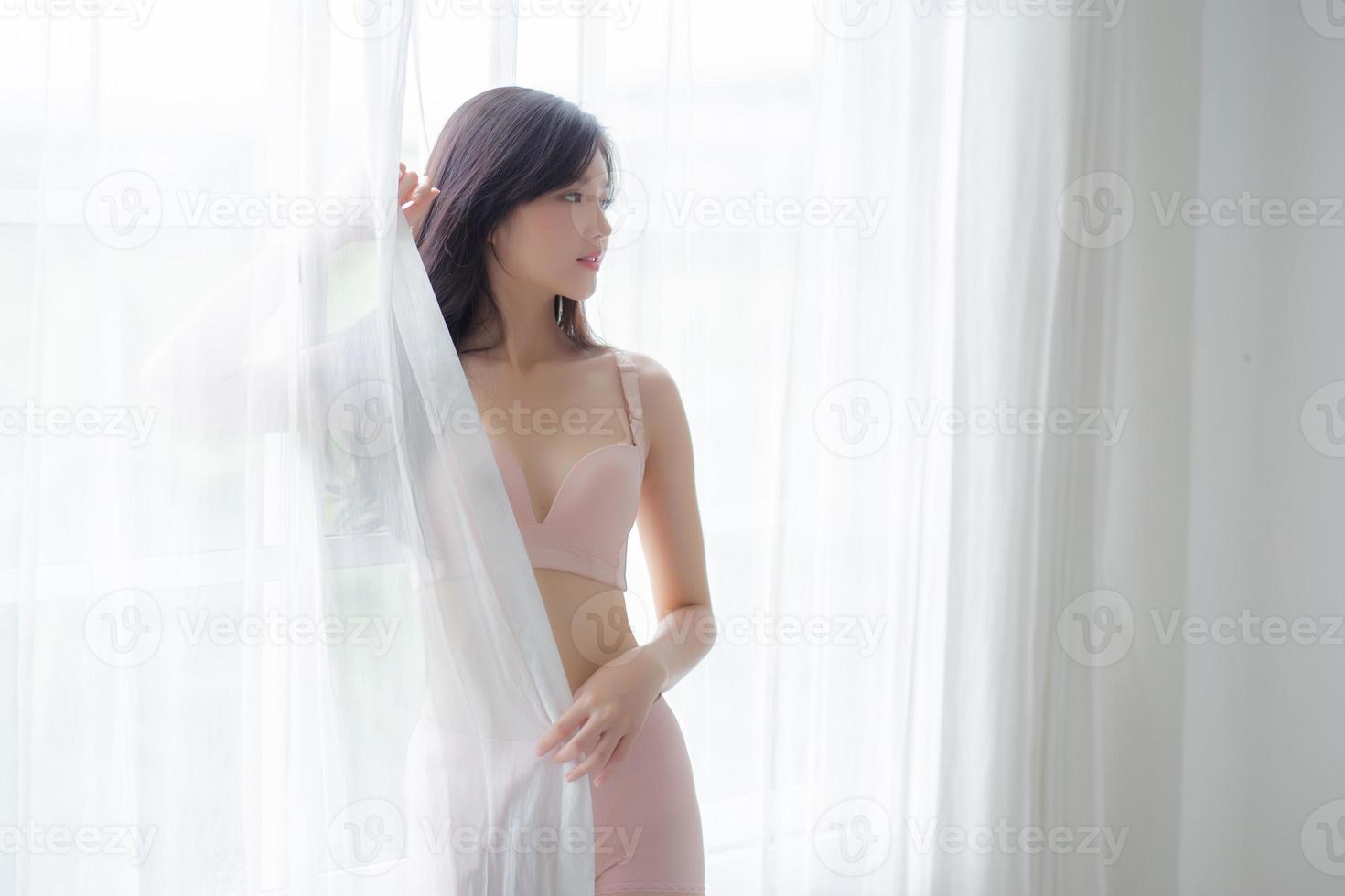 hermoso retrato joven mujer asiática sexy parada en la ventana y sonríe mientras se despierta con salud, cuerpo de niña feliz con frescura y alegre con bienestar, estilo de vida y concepto de relajación. foto