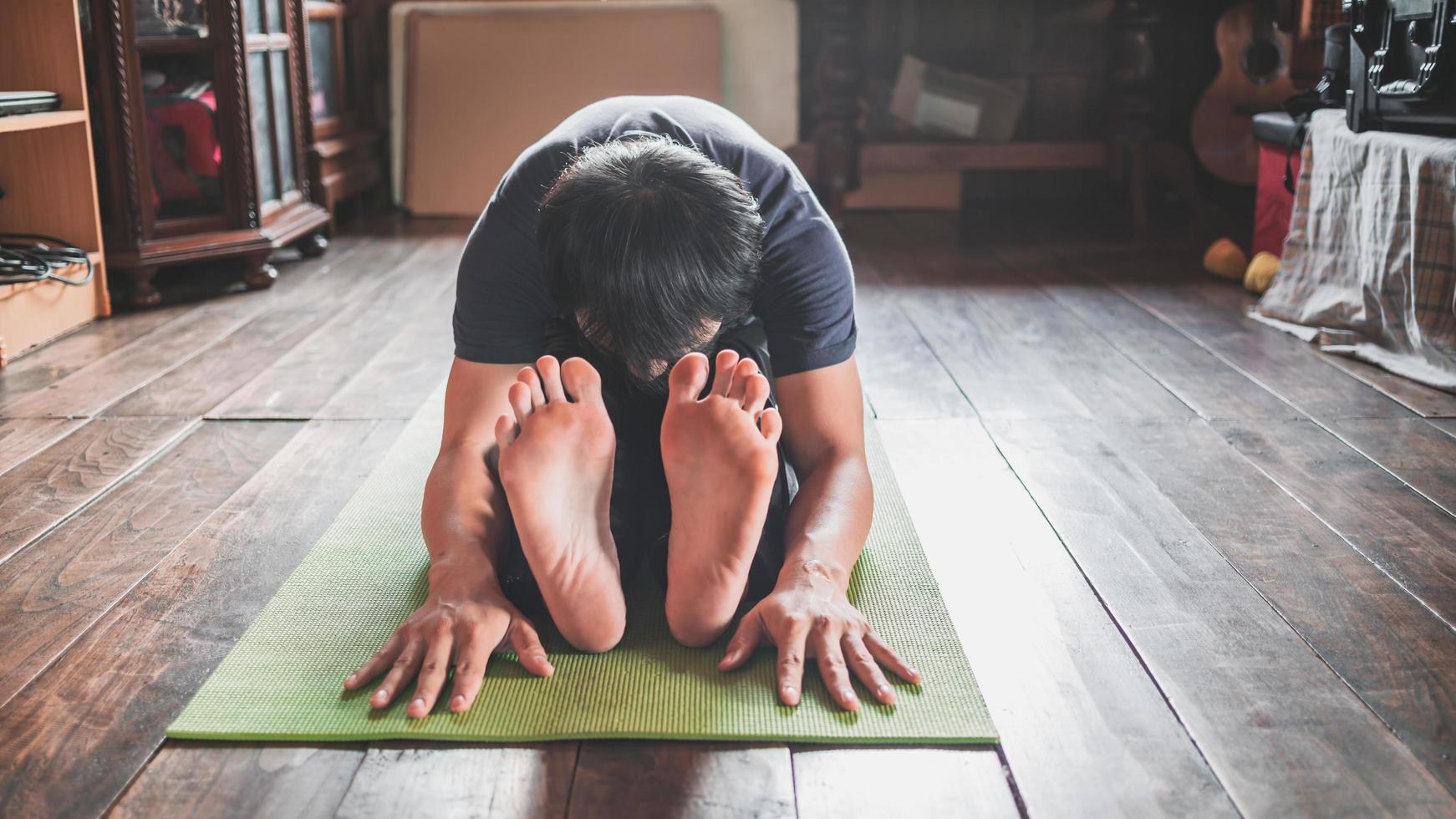 Young Asian man practicing yoga, sitting in Seated forward bend exercise, paschimottanasana pose on green mat yoga at wooden home. Healthy living photo