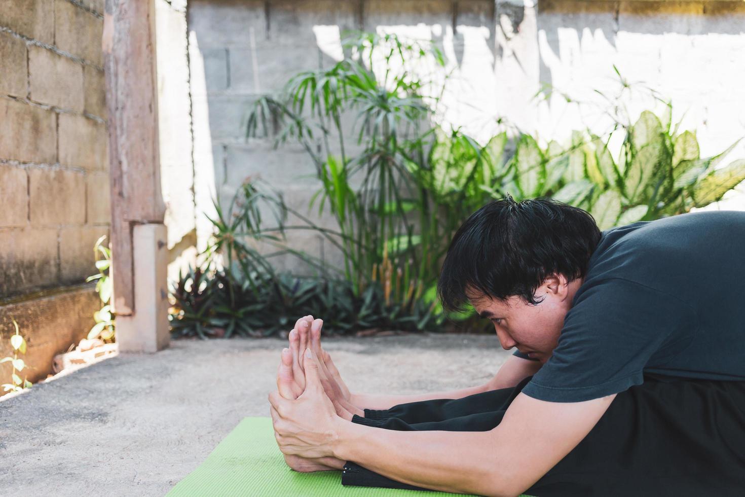 Young Asian man practicing yoga, sitting in Seated forward bend exercise, paschimottanasana pose on the green yoga mat, working out outdoor in the garden. healthy lifestyle and Healthy living concept photo