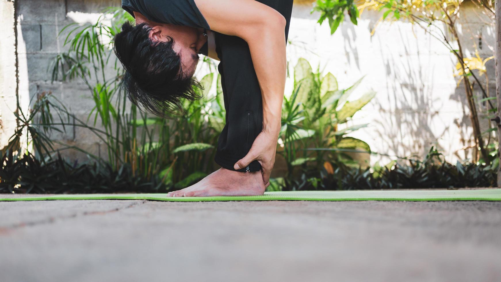 Healthy living concept of young Asian man practicing yoga asana Uttanasana - Standing Forward Fold pose, working out,  poses on a green yoga mat. outdoor exercise in the garden. healthy lifestyle photo
