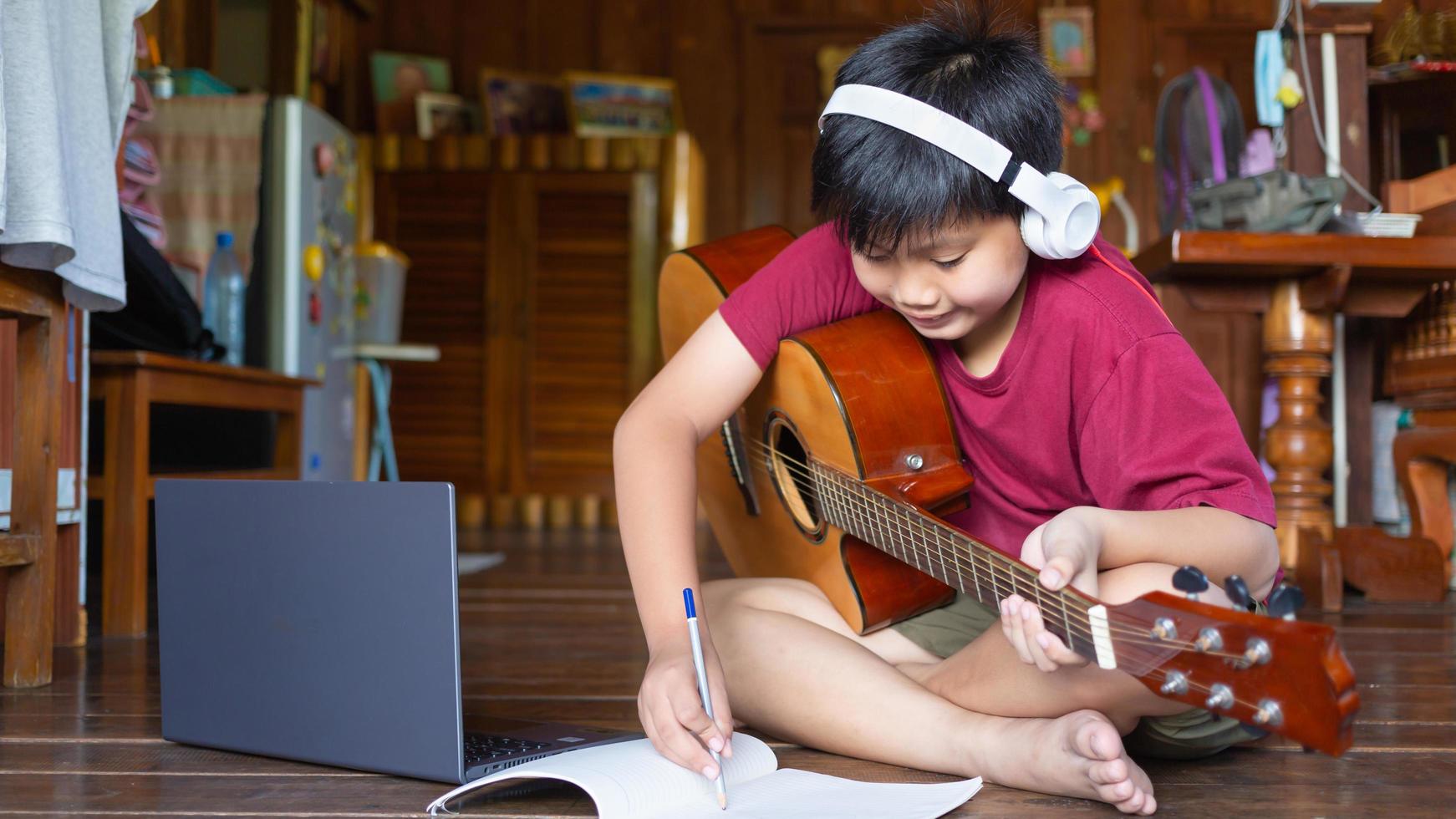 A cute Asian boy wearing headphones video calls learning  music and writing songs online on his computer laptop using an acoustic guitar happily at home kid creators concept photo