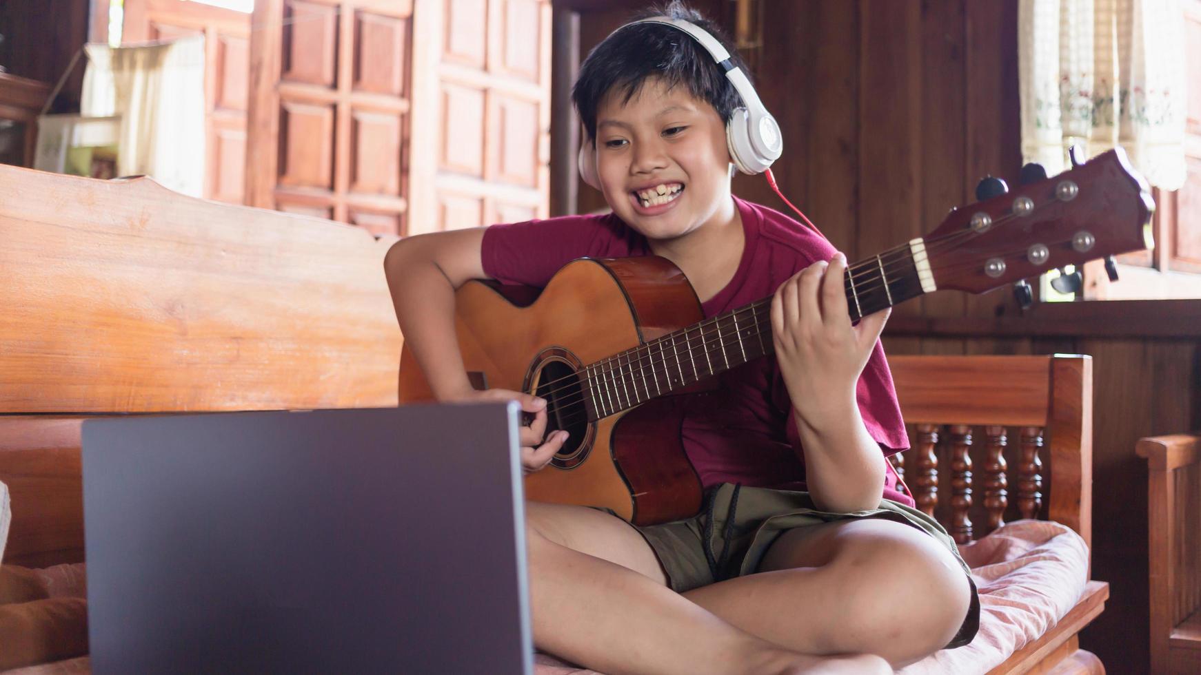 un lindo niño asiático con auriculares videollamadas aprendiendo música y escribiendo canciones en línea en su computadora portátil usando una guitarra acústica felizmente en casa concepto de creadores de niños foto