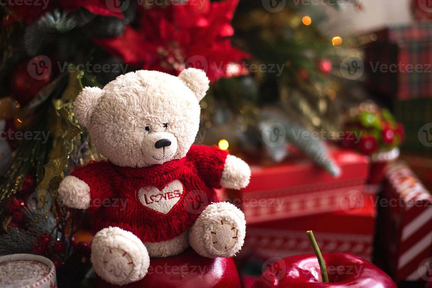 White Teddy bear in red knitted sweater with heart on the chest and the words Love near Christmas tree among the gift boxes. Gift to your beloved, Declaration of love, poinsettia decor. Copy space photo