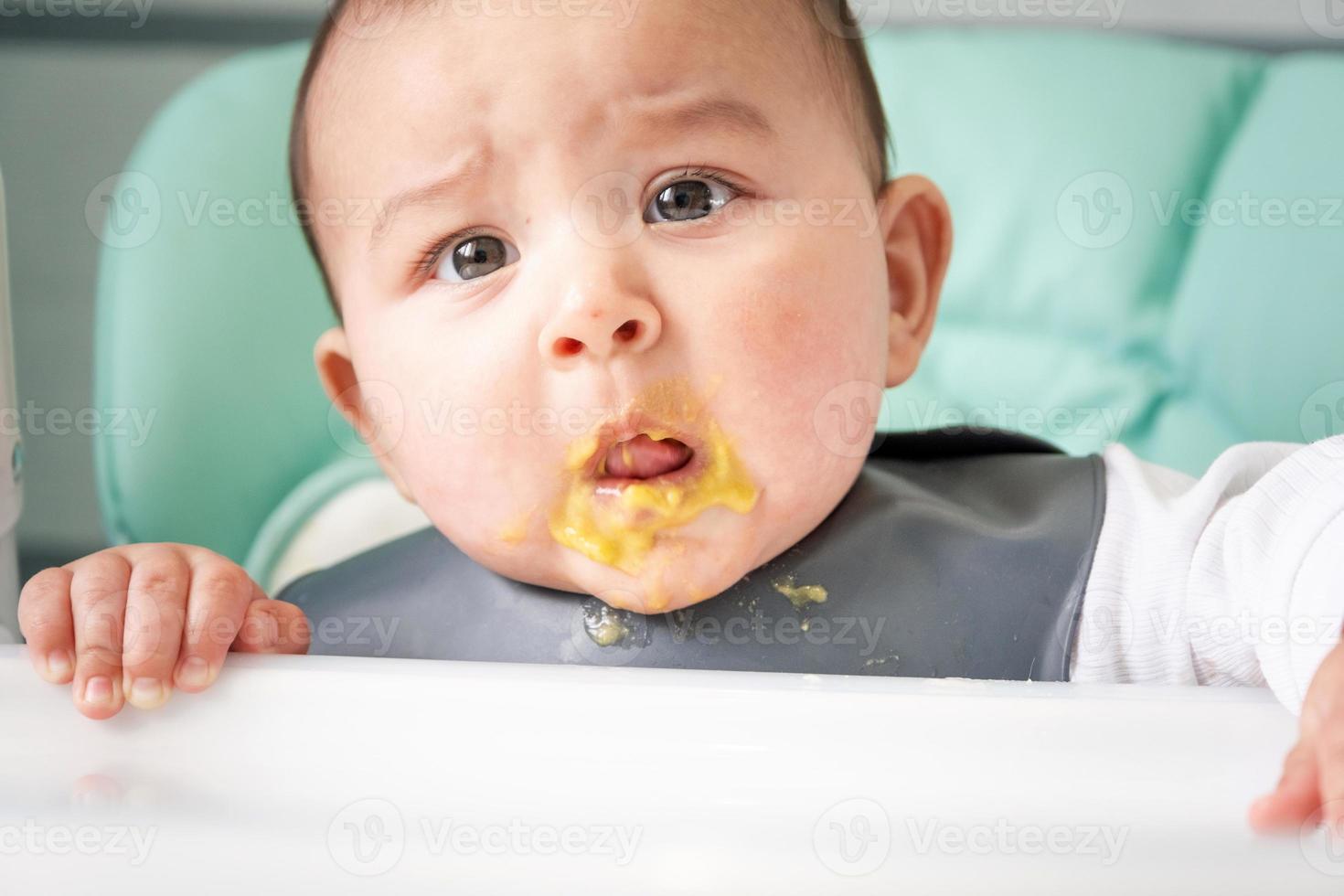 A dirty surprised baby stained in vegetable puree at the feeding table. Introduction of complementary foods, do not like the taste, new food. Close-up, portrait photo