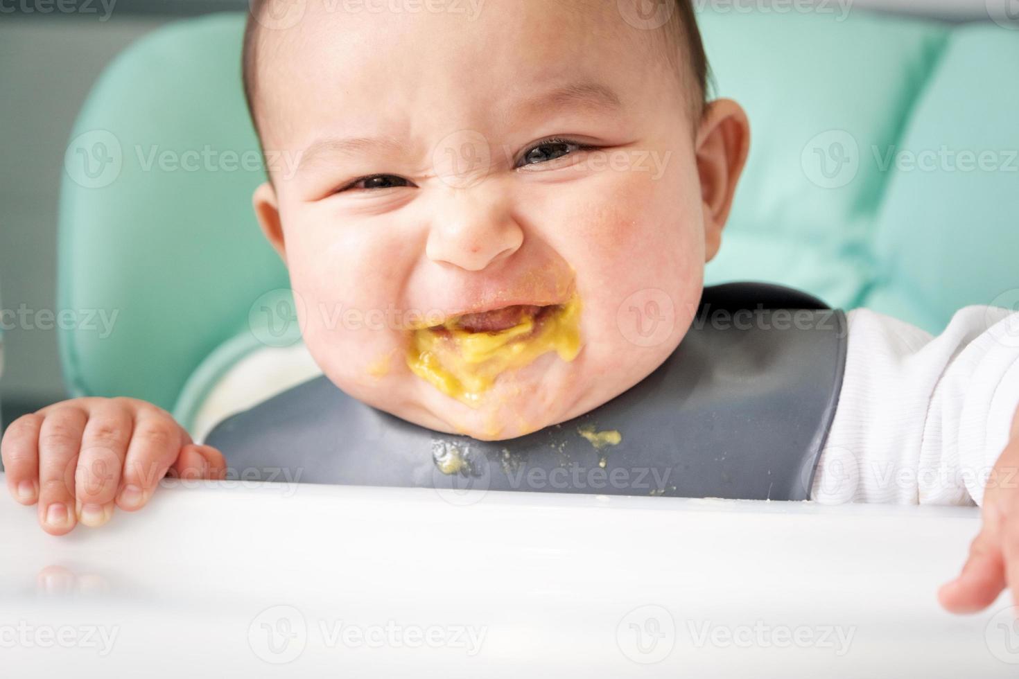 A dirty, joyful baby at the table on a feeding chair got his mouth dirty in vegetable puree. Introduction of complementary foods, the child likes to eat. Portrait close-up photo