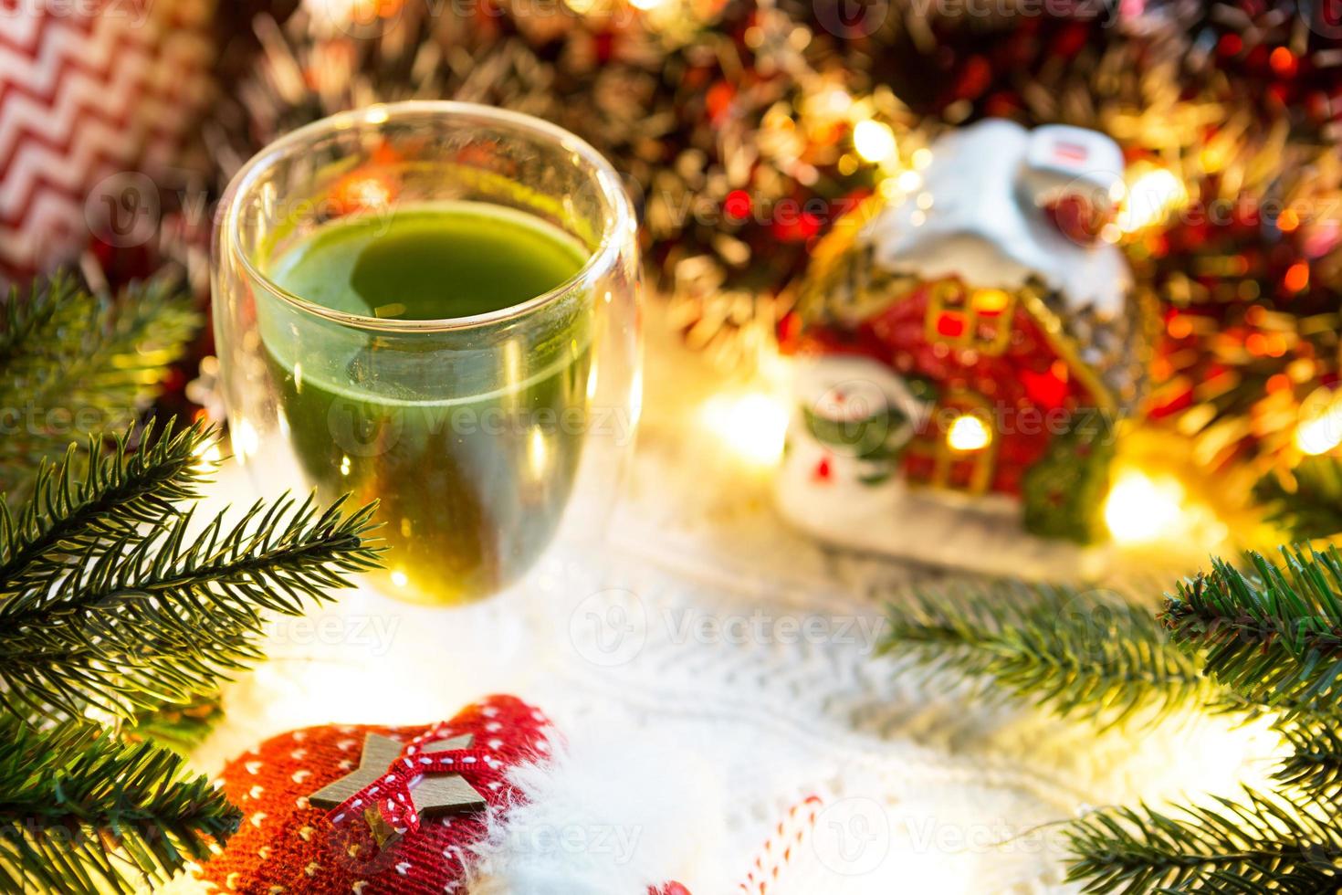 vaso transparente de vidrio de doble pared con té matcha japonés en la mesa con decoración navideña. atmósfera de año nuevo, guirnaldas y oropel, rama de abeto, acogedora manta de punto, pelota, servilleta a rayas foto