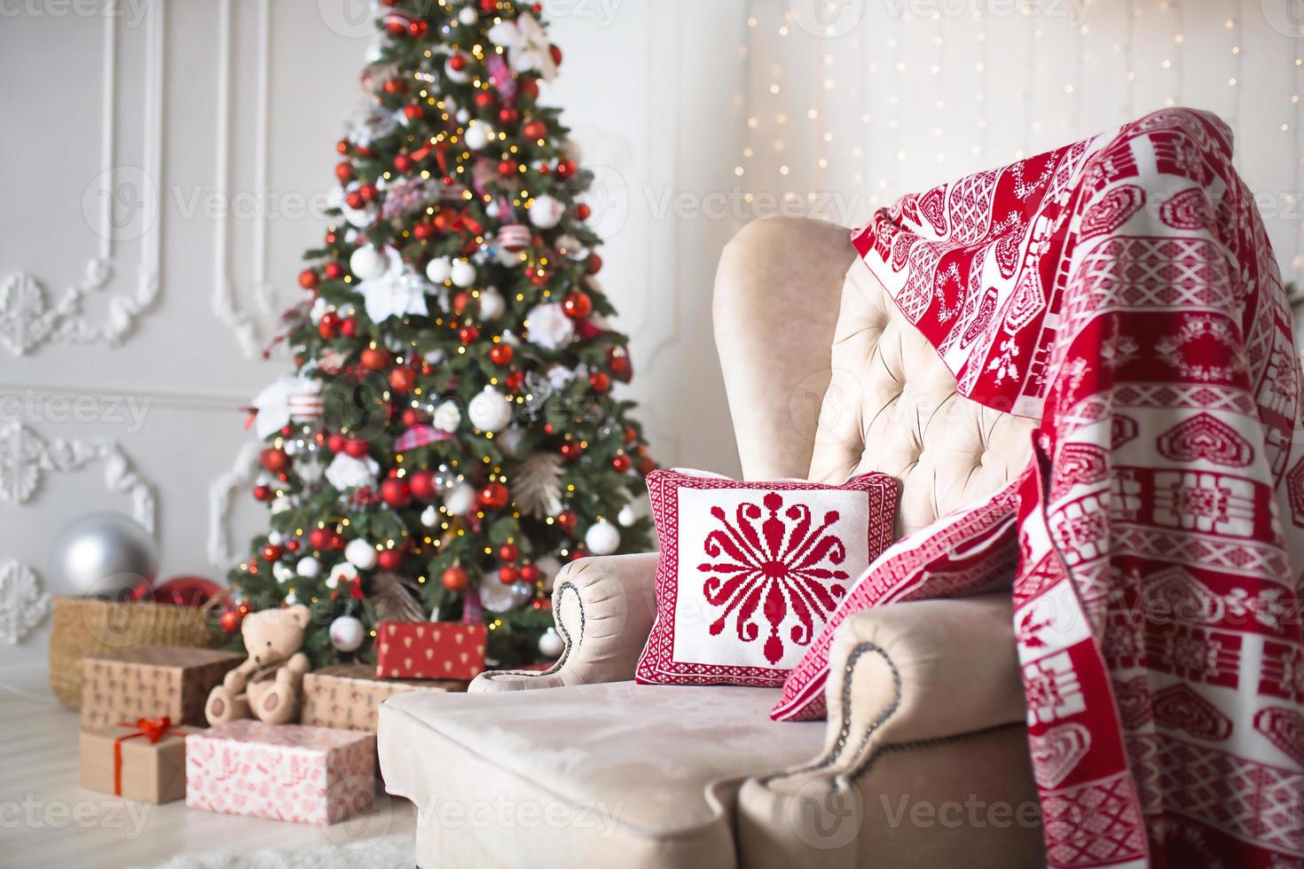 Christmas tree with red and white decor in a white living room with gifts in boxes, a chair with pillows and a blanket with winter ornaments, a fireplace, a fur rug. New year, European style. photo