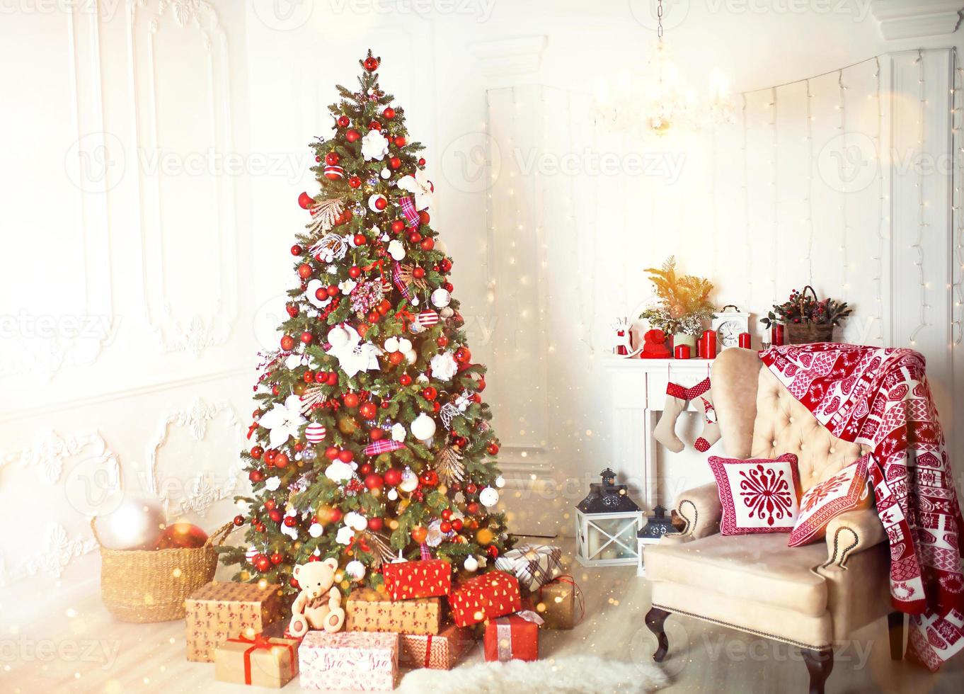 Christmas tree with red and white decor in a white living room with gifts in boxes, a chair with pillows and a blanket with winter ornaments, a fireplace, a fur rug. New year, European style. photo