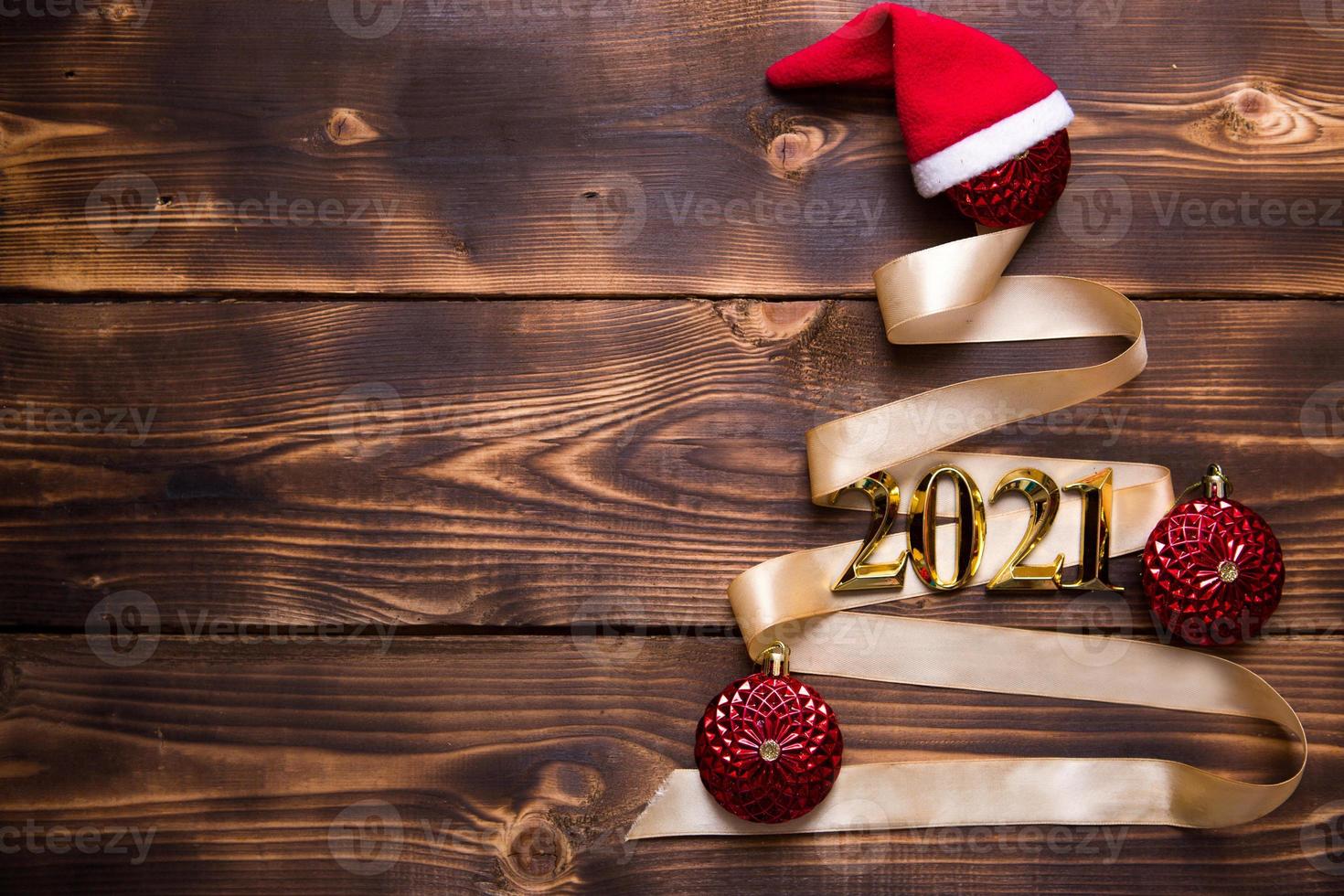 A Christmas tree made of gold ribbon with the numbers 2021 is decorated with red balloons on a dark wooden background. Flat lay. Space for text. New year, Santa hat. photo