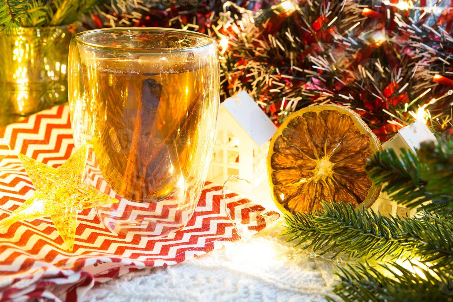 vaso de vidrio transparente de doble pared con té caliente y palitos de canela en la mesa con decoración navideña y casa pequeña. ambiente de año nuevo, rodaja de naranja seca, guirnalda, rama de abeto, acogedor foto