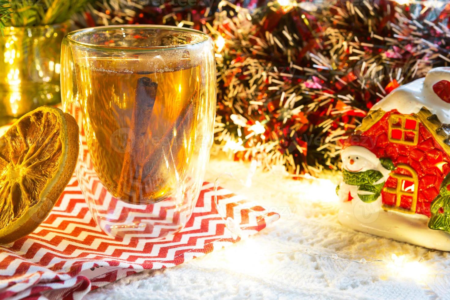 vaso de vidrio transparente de doble pared con té caliente y palitos de canela en la mesa con decoración navideña y casa pequeña. ambiente de año nuevo, rodaja de naranja seca, guirnalda, rama de abeto, acogedor foto