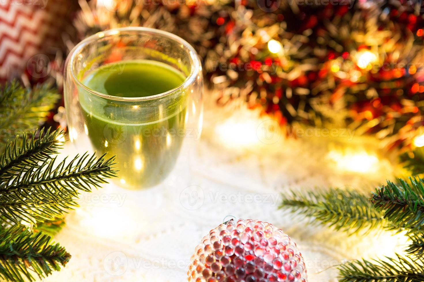 vaso transparente de vidrio de doble pared con té matcha japonés en la mesa con decoración navideña. atmósfera de año nuevo, guirnaldas y oropel, rama de abeto, acogedora manta de punto, pelota, servilleta a rayas foto