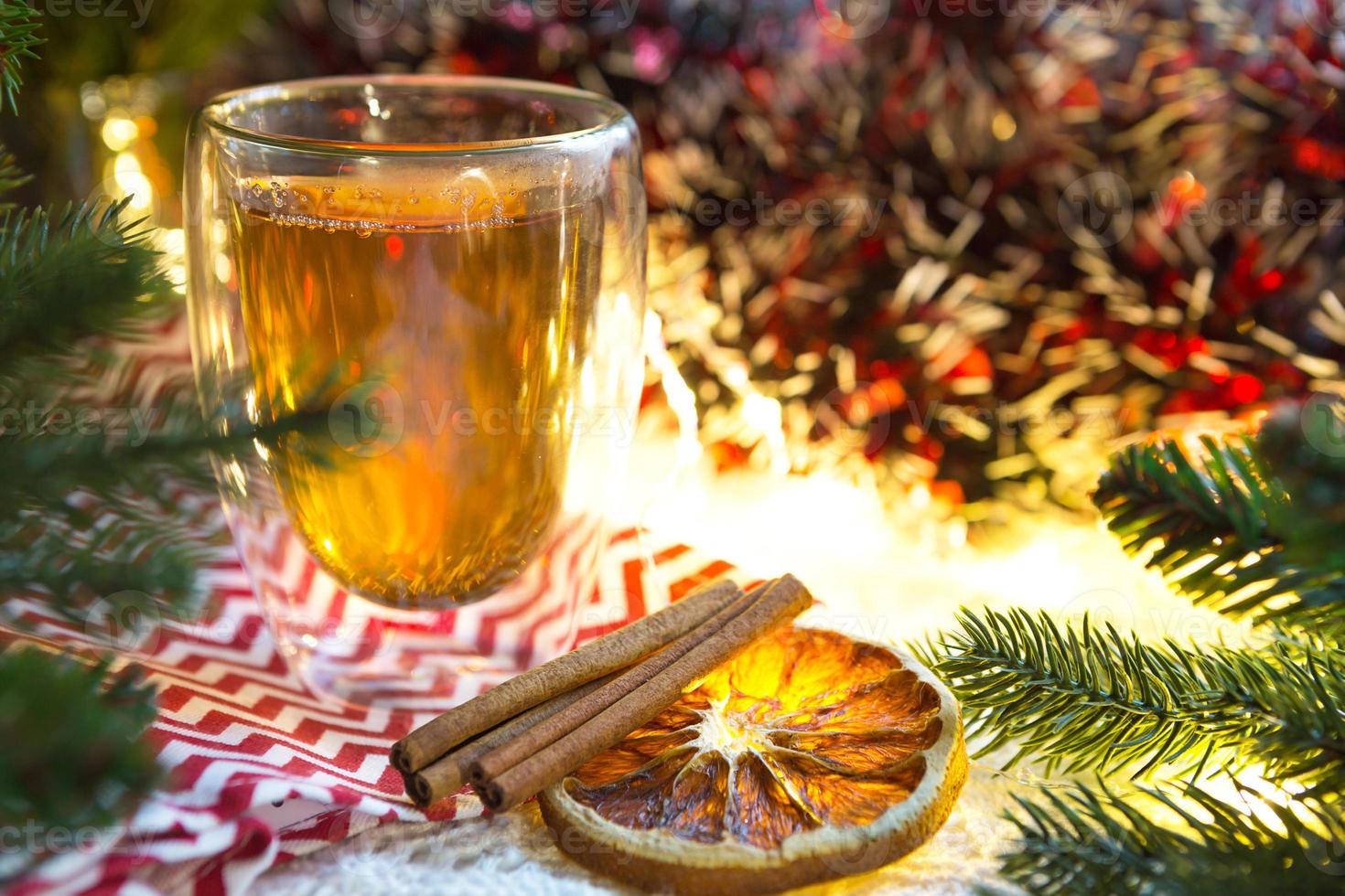 Transparent double-walled glass tumbler with hot tea and cinnamon sticks on the table with Christmas decor. New year's atmosphere, slice of dried orange, garland and tinsel, spruce branch, cozy photo