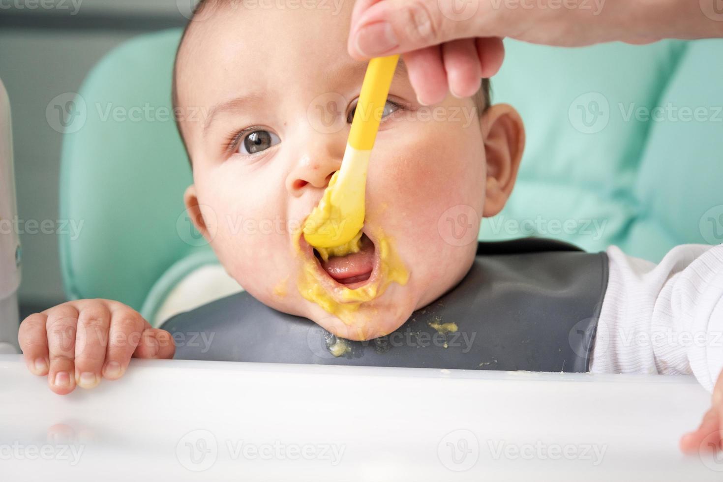 Mom feeds the baby with a spoon of vegetable puree at the children's feeding table. Baby's appetite, healthy nutrition, introduction of complementary foods. Copyspace, mock up photo
