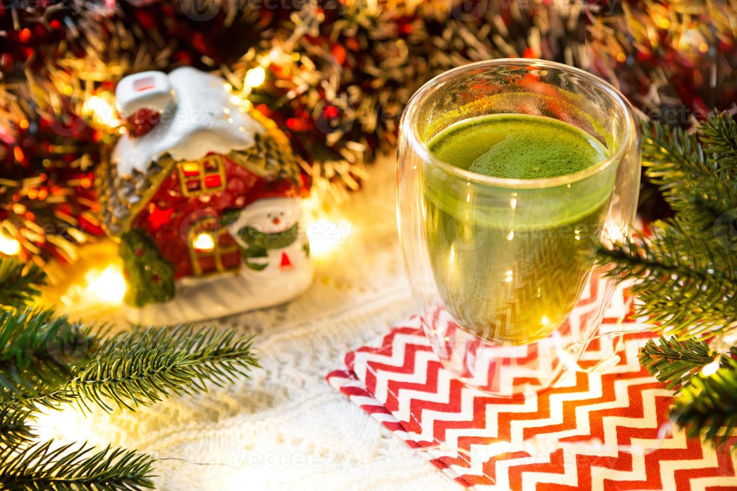 Transparent double-walled glass tumbler with Japanese matcha tea on table with Christmas decor. New year's atmosphere, garland and tinsel, spruce branch, cozy, knitted blanket, ball, striped napkin photo