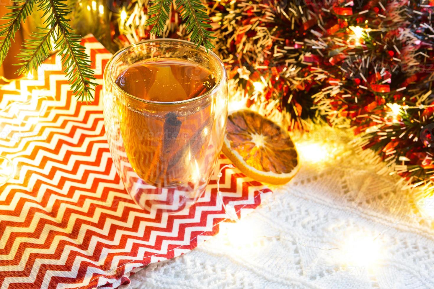 Transparent double-walled glass tumbler with hot tea and cinnamon sticks on the table with Christmas decor. New year's atmosphere, slice of dried orange, garland and tinsel, spruce branch, cozy photo