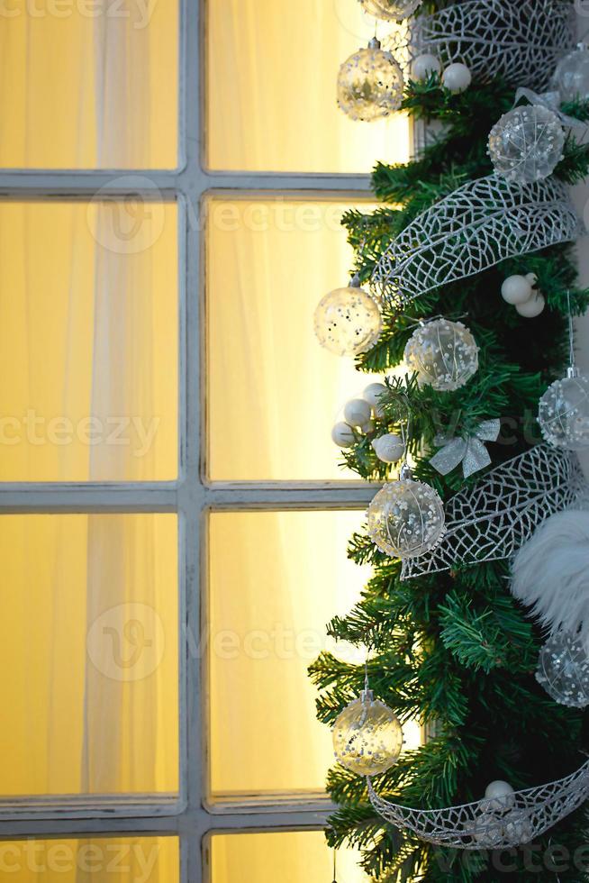 Christmas decorations made of spruce branches and white transparent balls and ribbons frame the window with wooden frame and yellow light. New year festive atmosphere, comfort of home. Space for text photo