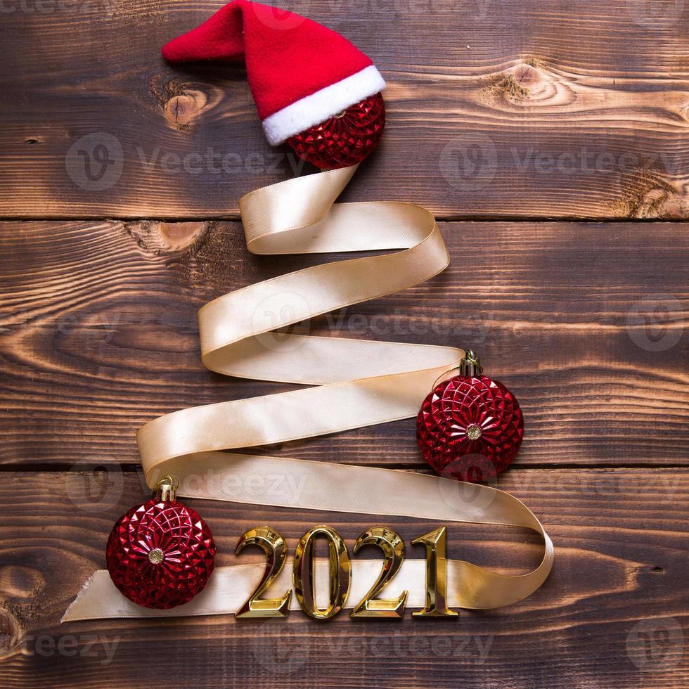 A Christmas tree made of gold ribbon with the numbers 2021 is decorated with red balloons on a dark wooden background. Flat lay. Space for text. New year, Santa hat. photo