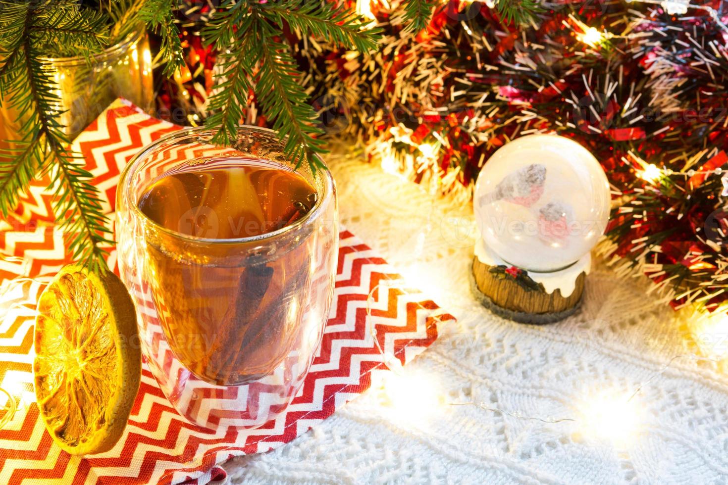 vaso transparente de vidrio de doble pared con té caliente y palitos de canela en la mesa con decoración navideña. ambiente de año nuevo, rodaja de naranja seca, guirnalda y oropel, globo de nieve con camachuelos foto