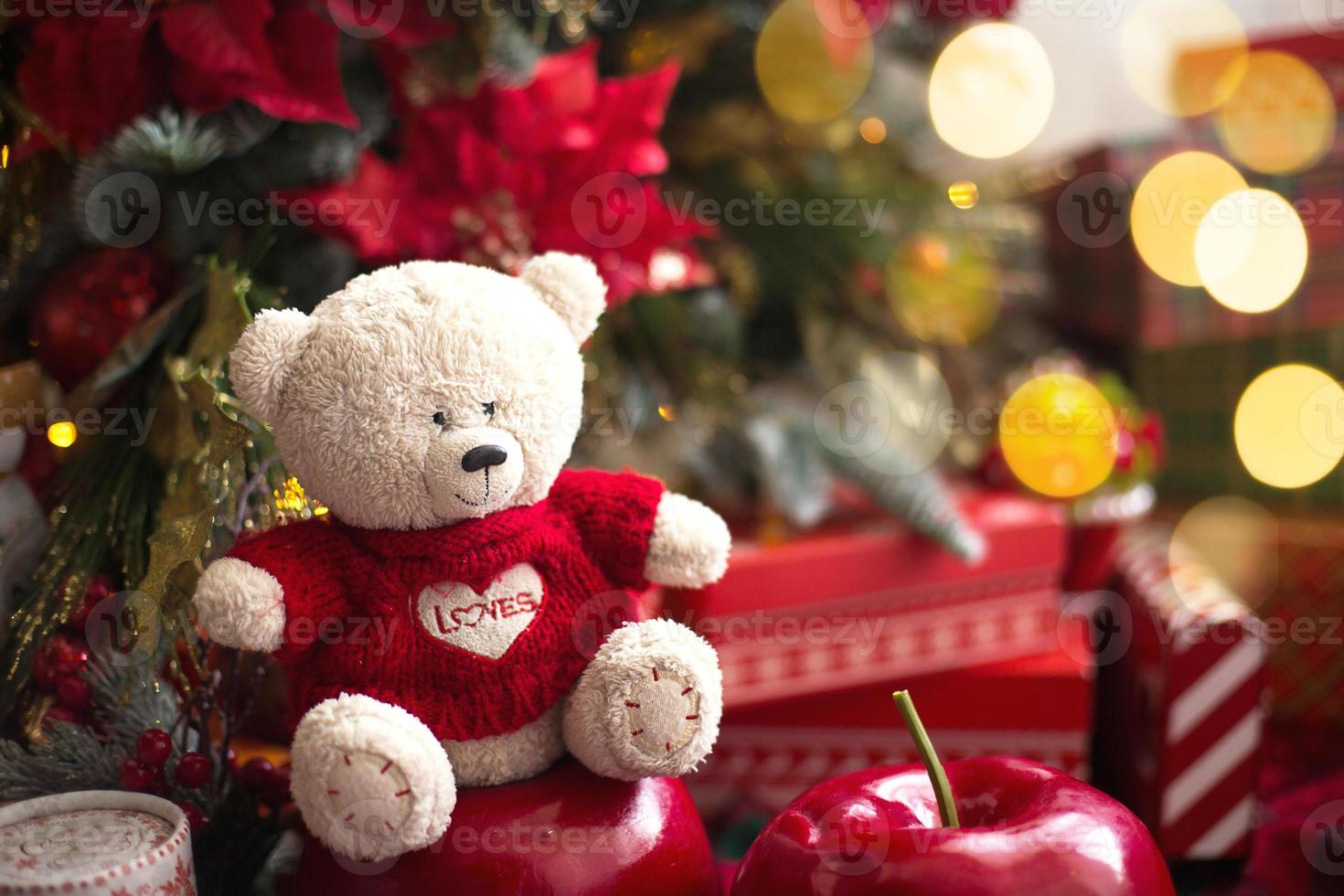 White Teddy bear in red knitted sweater with heart on the chest and the words Love near Christmas tree among the gift boxes. Gift to your beloved, Declaration of love, poinsettia decor. Copy space photo