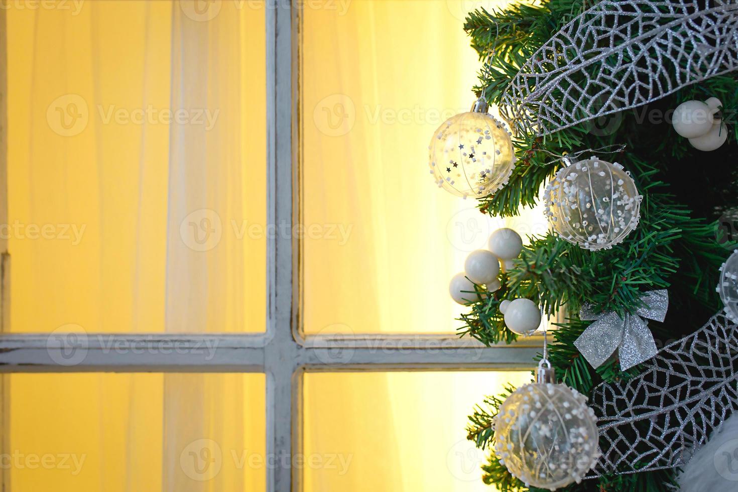 adornos navideños hechos de ramas de abeto y bolas y cintas transparentes blancas enmarcan la ventana con marco de madera y luz amarilla. ambiente festivo de año nuevo, comodidad del hogar. espacio para texto foto