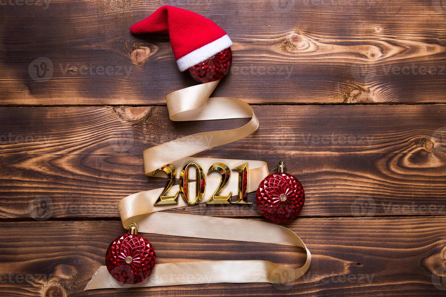 A Christmas tree made of gold ribbon with the numbers 2021 is decorated with red balloons on a dark wooden background. Flat lay. Space for text. New year, Santa hat. photo