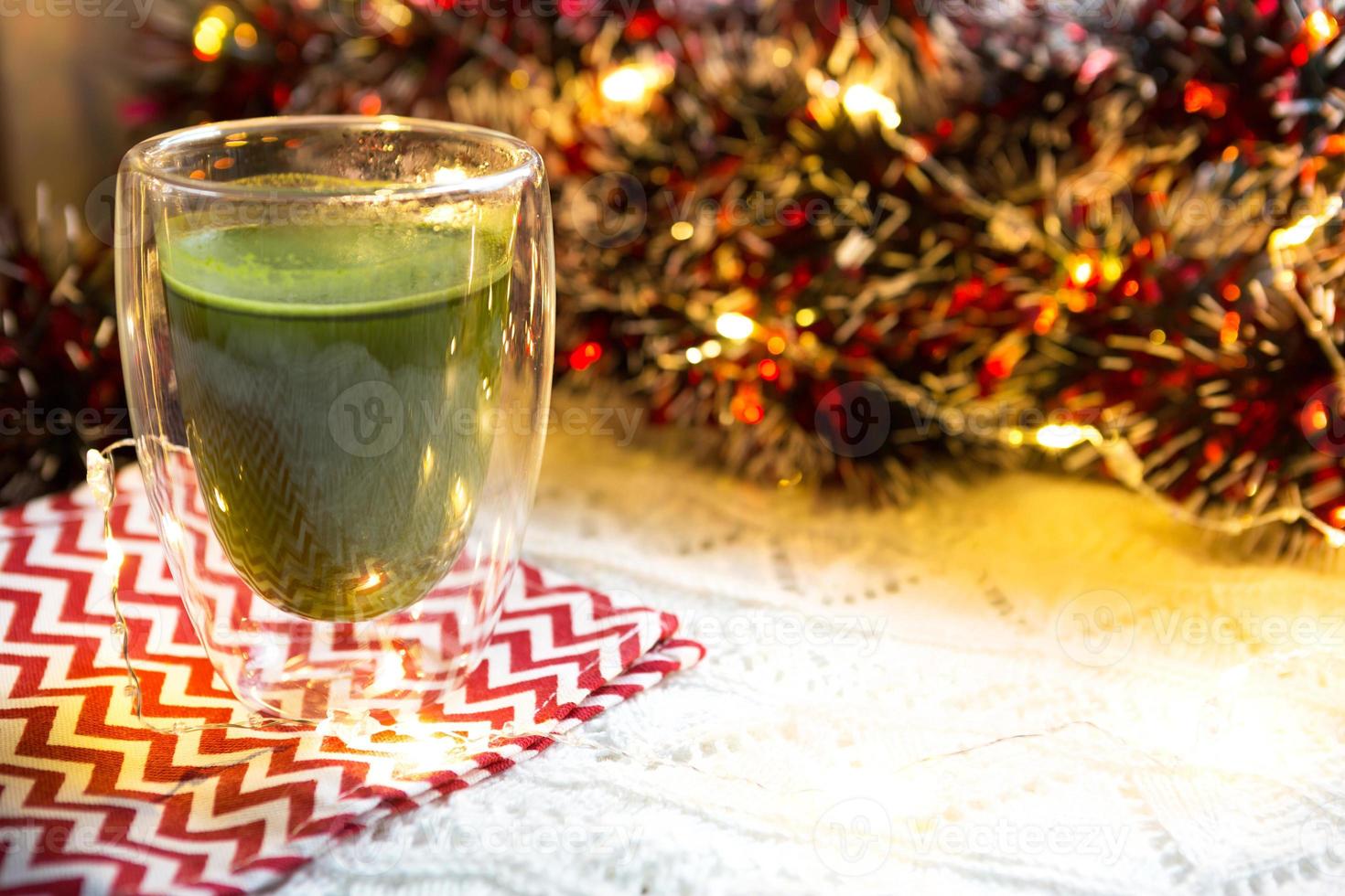 vaso transparente de vidrio de doble pared con té matcha japonés en la mesa con decoración navideña. atmósfera de año nuevo, guirnaldas y oropel, rama de abeto, acogedora manta de punto, pelota, servilleta a rayas foto