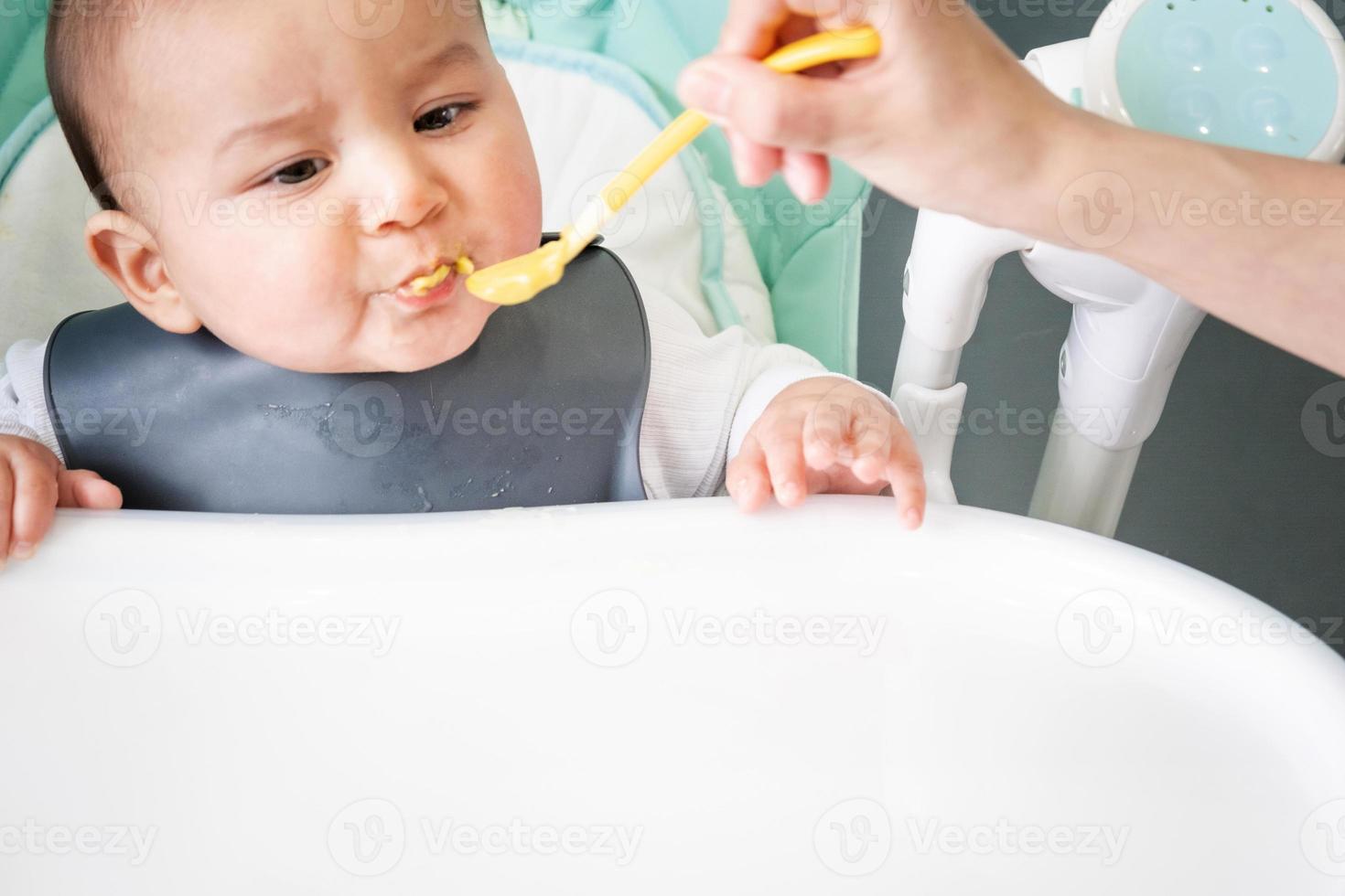 Mom feeds the baby with a spoon of vegetable puree at the children's feeding table. Baby's appetite, healthy nutrition, introduction of complementary foods. Copyspace, mock up photo