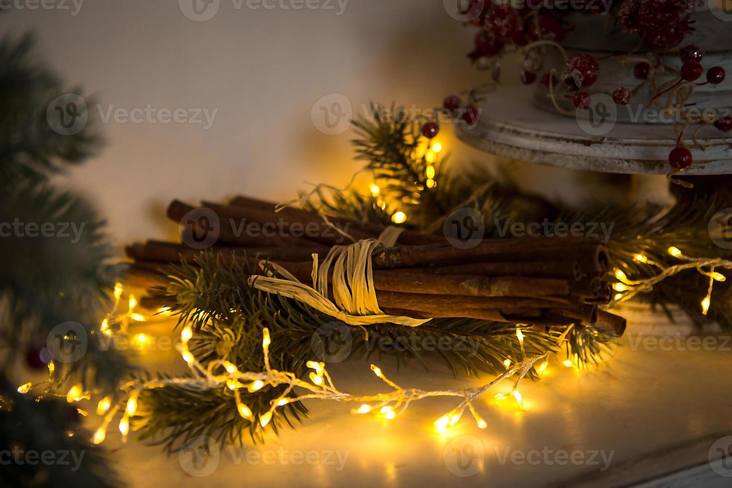 Cinnamon sticks tied with string in yellow lights garlands on Christmas tree branches and red berries. The atmosphere of the new year festive background, details of decorations photo