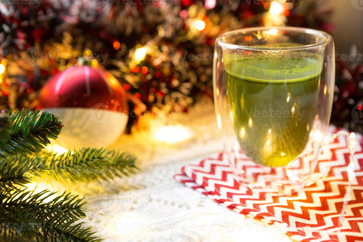 vaso transparente de vidrio de doble pared con té matcha japonés en la mesa con decoración navideña. atmósfera de año nuevo, guirnaldas y oropel, rama de abeto, acogedora manta de punto, pelota, servilleta a rayas foto