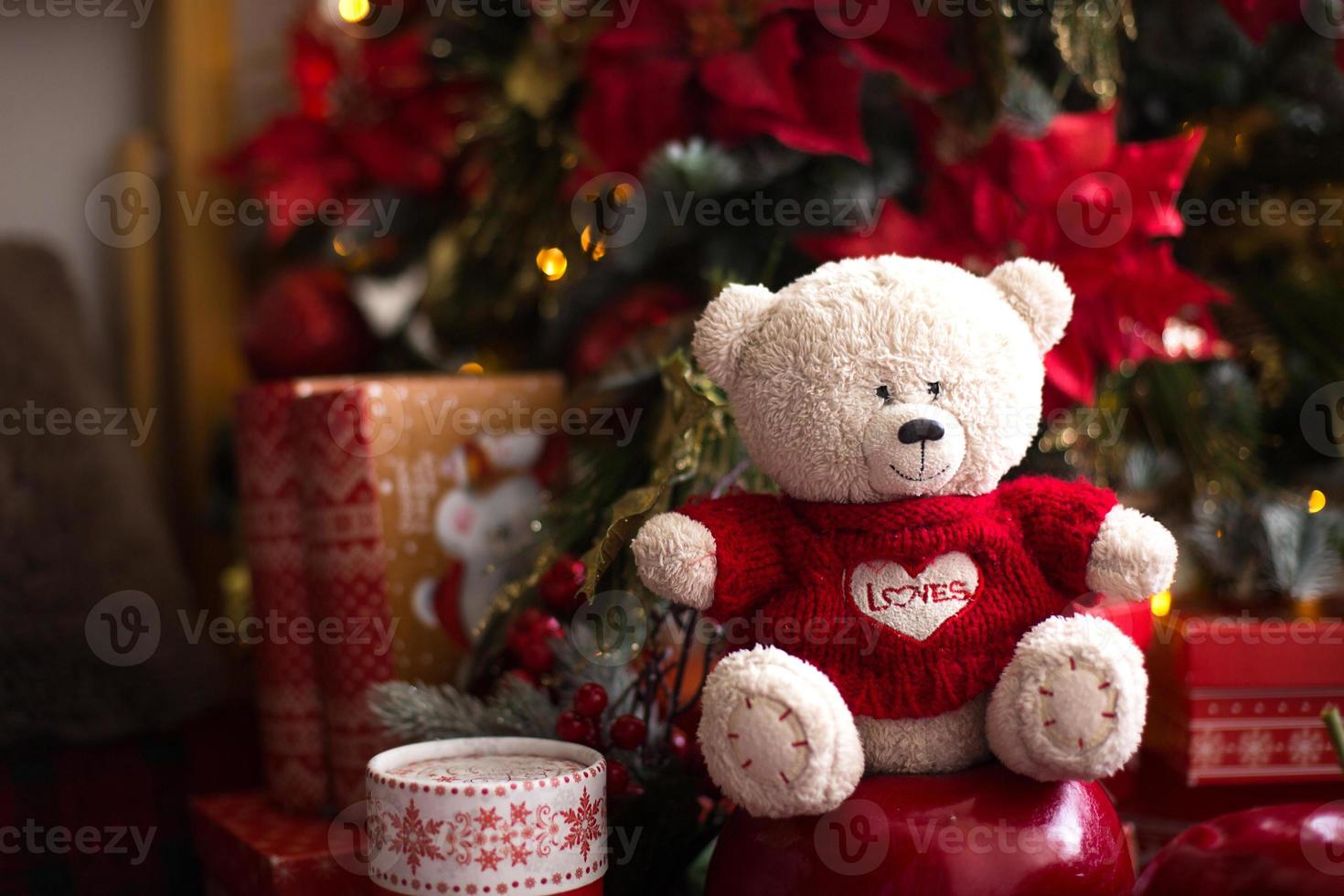 oso de peluche blanco con suéter de punto rojo con corazón en el pecho y las palabras amor cerca del árbol de navidad entre las cajas de regalo. regalo para tu amada, declaración de amor, decoración de poinsettia. copie el espacio foto