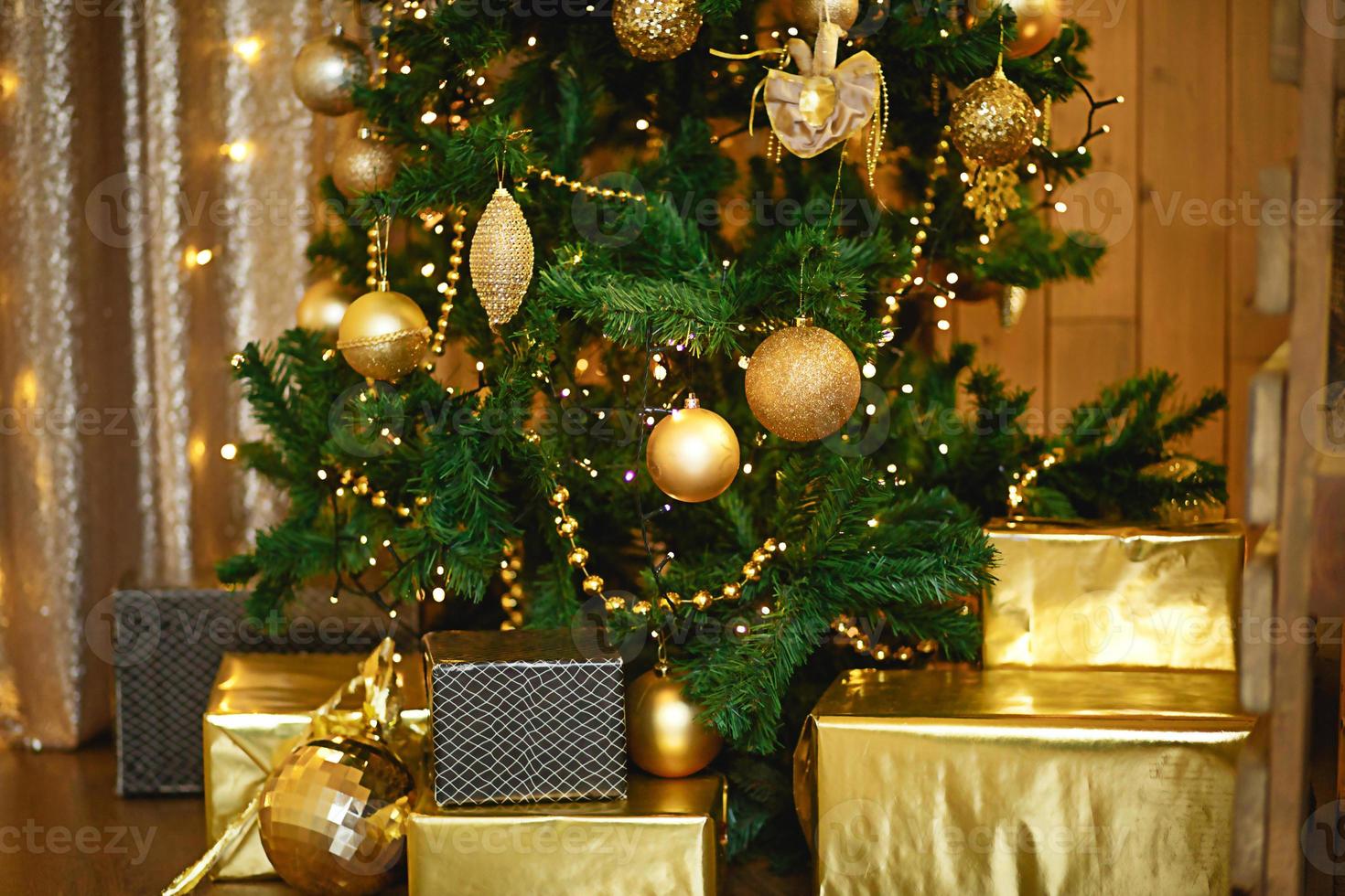 Christmas green artificial fir tree in the Golden room with lights garlands decorated with Golden balls, beads and toys. The box with the gifts in foil and black paper. New Year. Space for text photo