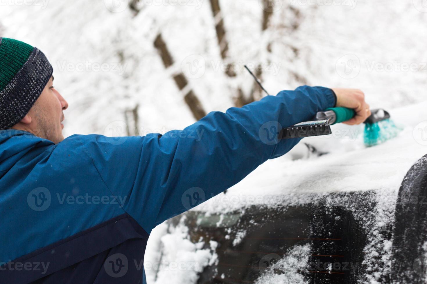 A man brushes snow from a car after a snowfall. A hand in a blue jacket with a car broom on the white body. Winter weather conditions photo