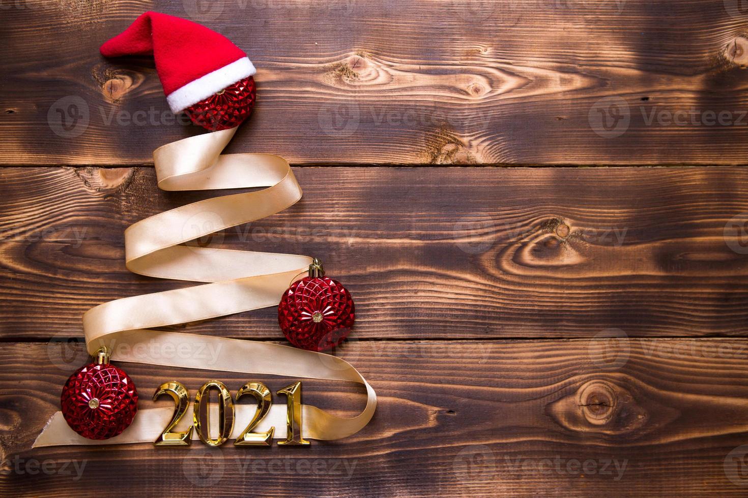 A Christmas tree made of gold ribbon with the numbers 2021 is decorated with red balloons on a dark wooden background. Flat lay. Space for text. New year, Santa hat. photo