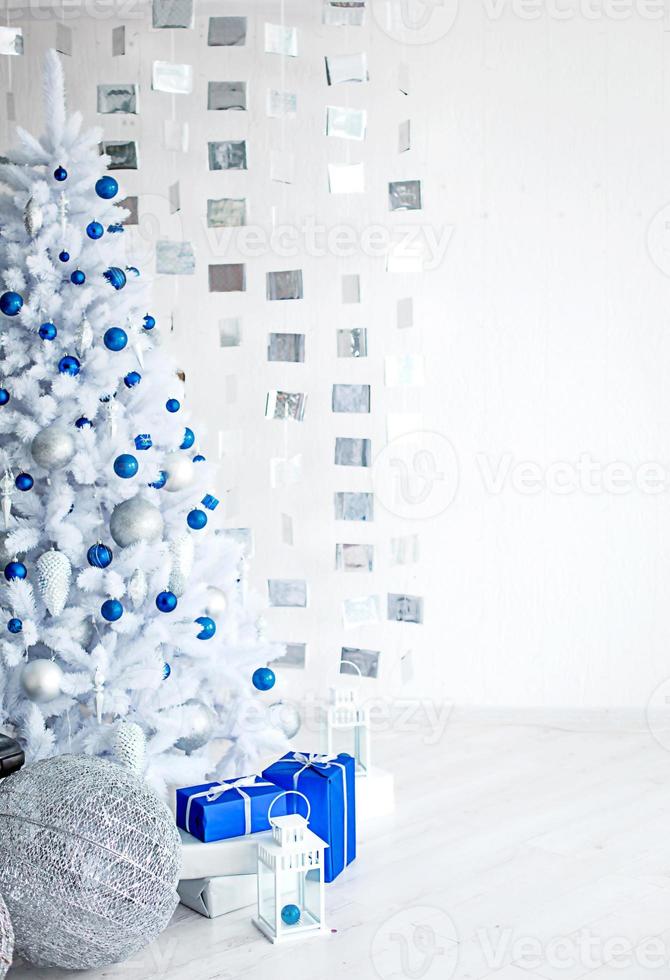 Christmas white artificial fir tree in a light gray room is decorated with blue and silver balloons. Boxes with gifts, decorations made of cubes on the wall. New Year. Space for text, cold background photo