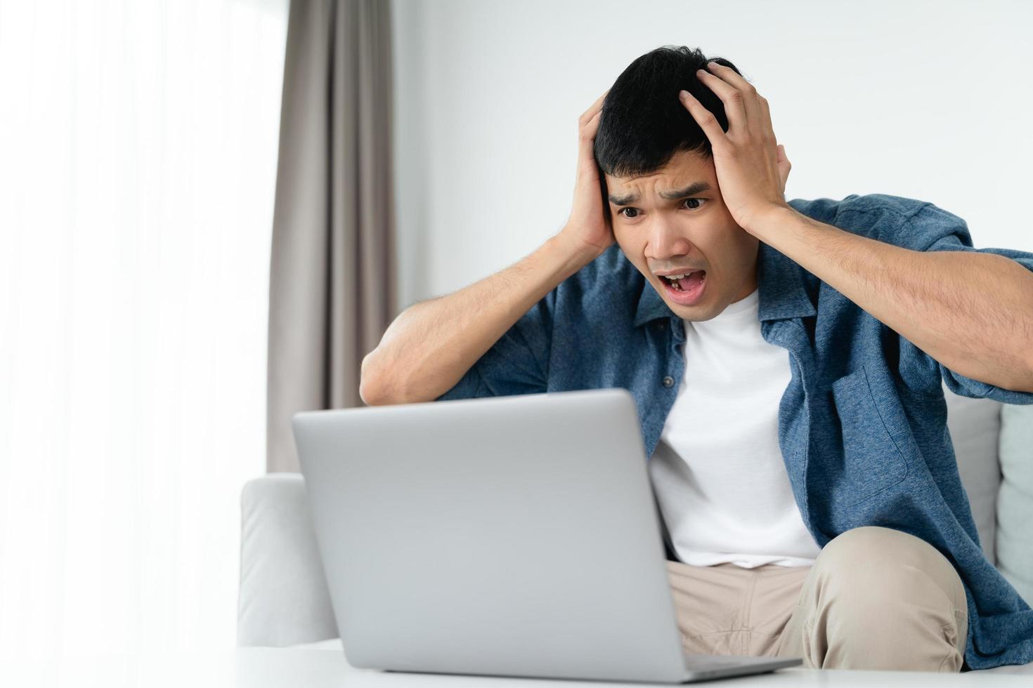 Shocked surprised Asian man looking at laptop computer screen sitting in the living room. photo