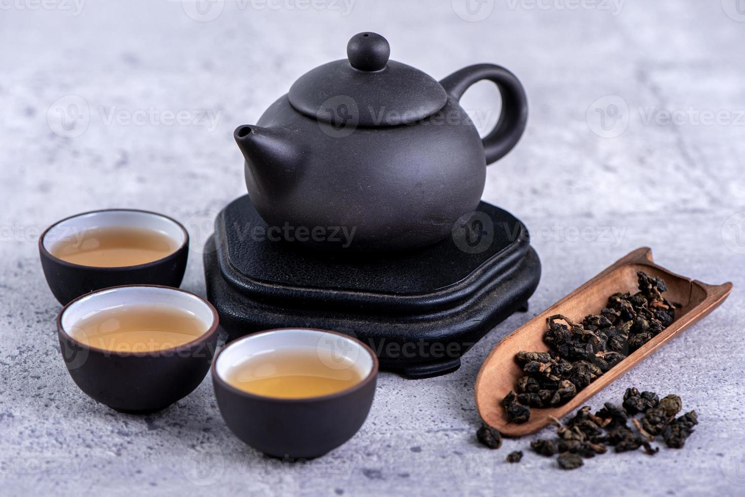 Hot tea in black teapot and cups and dry tea leaves over bright gray cement background, close up, copy space design concept. photo