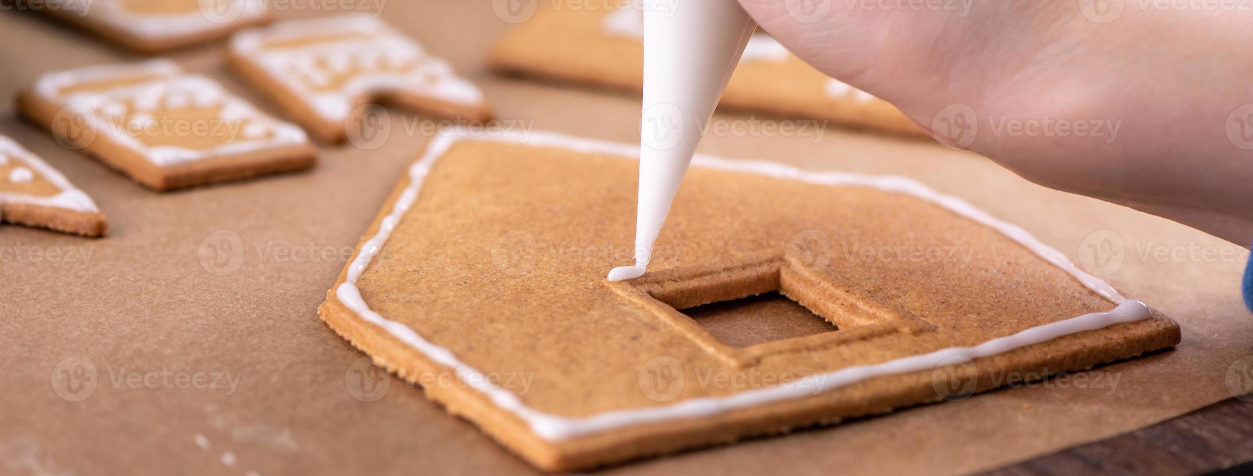 Young woman is decorating Christmas Gingerbread House cookies biscuit at home with frosting topping in icing bag, close up, lifestyle. photo