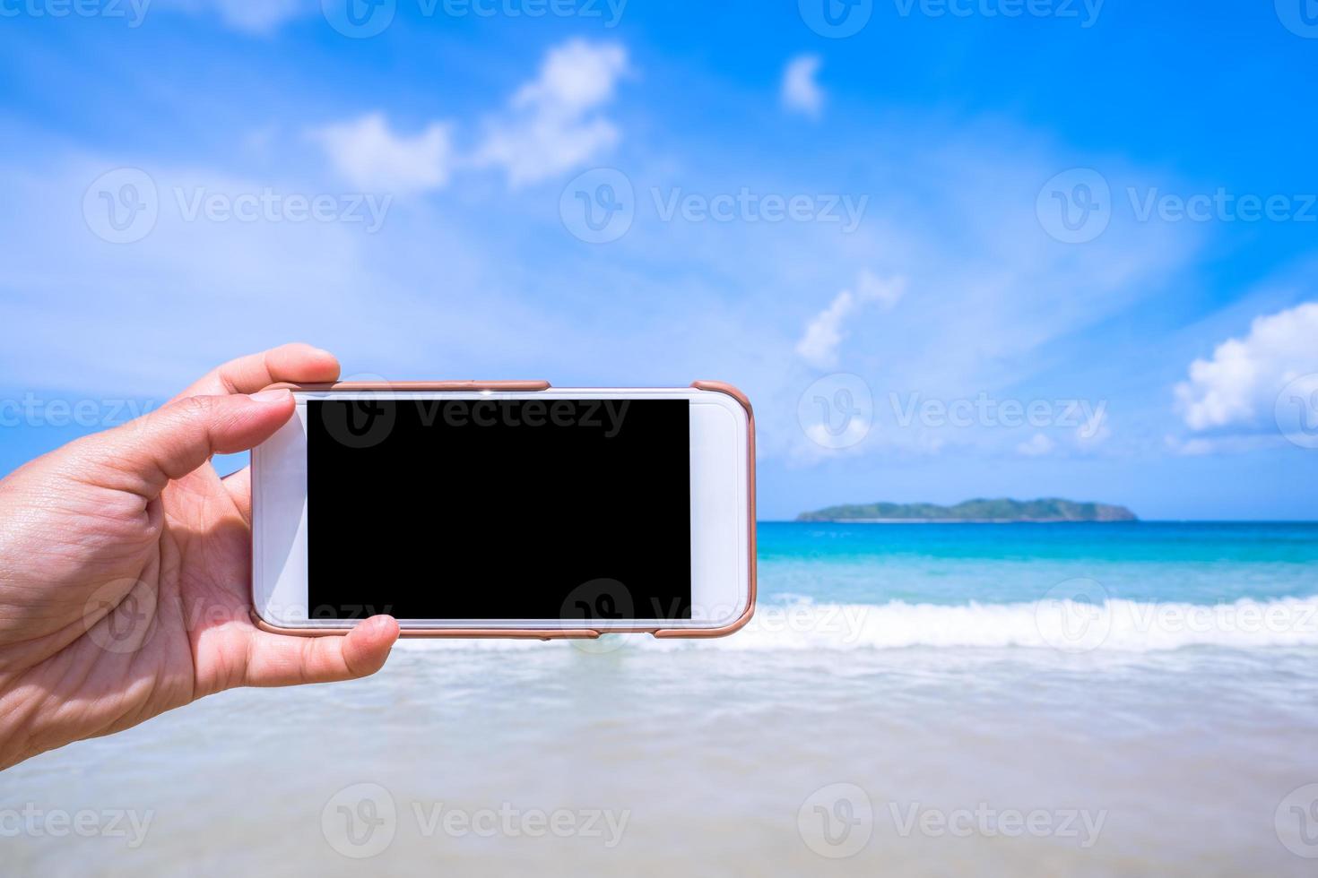 Tourist using phone at the beach with the sea, hand holding white mobile smart phone smartphone, travel working concept, blurry background, close up. photo