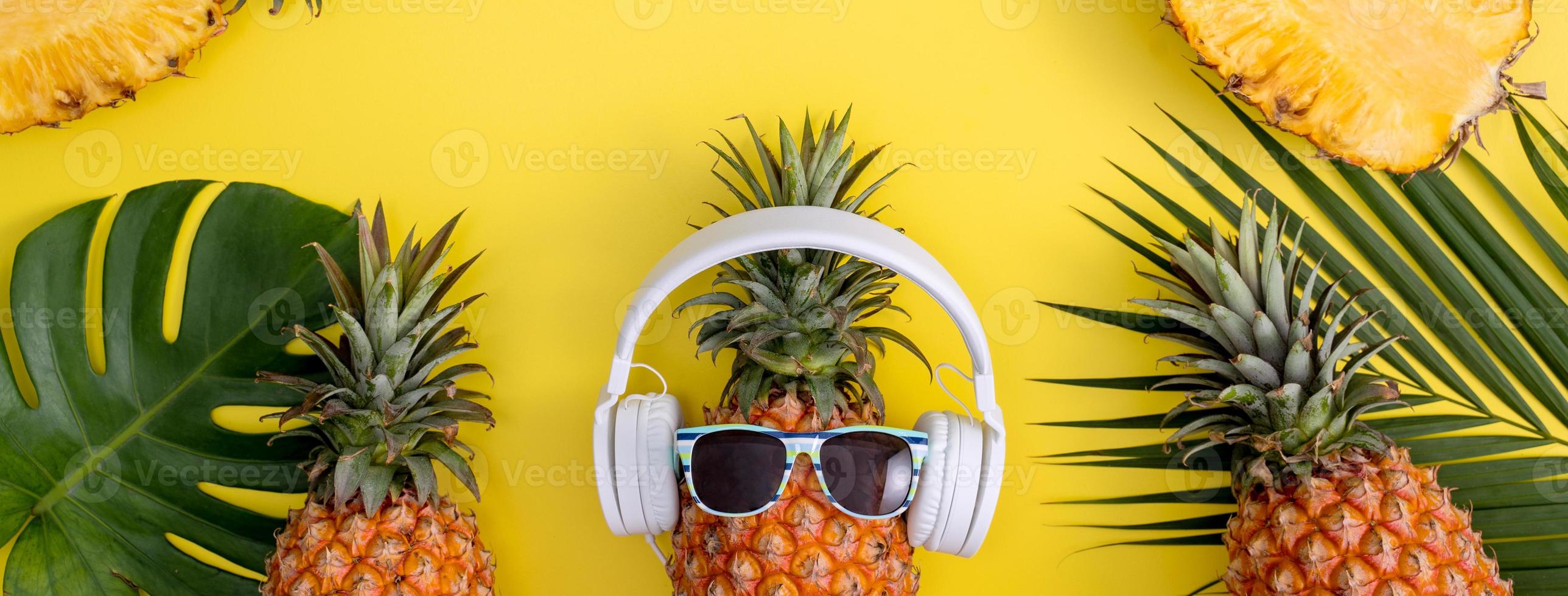 Funny pineapple wearing white headphone, listen music, isolated on yellow background with tropical palm leaves, top view, flat lay design concept. photo