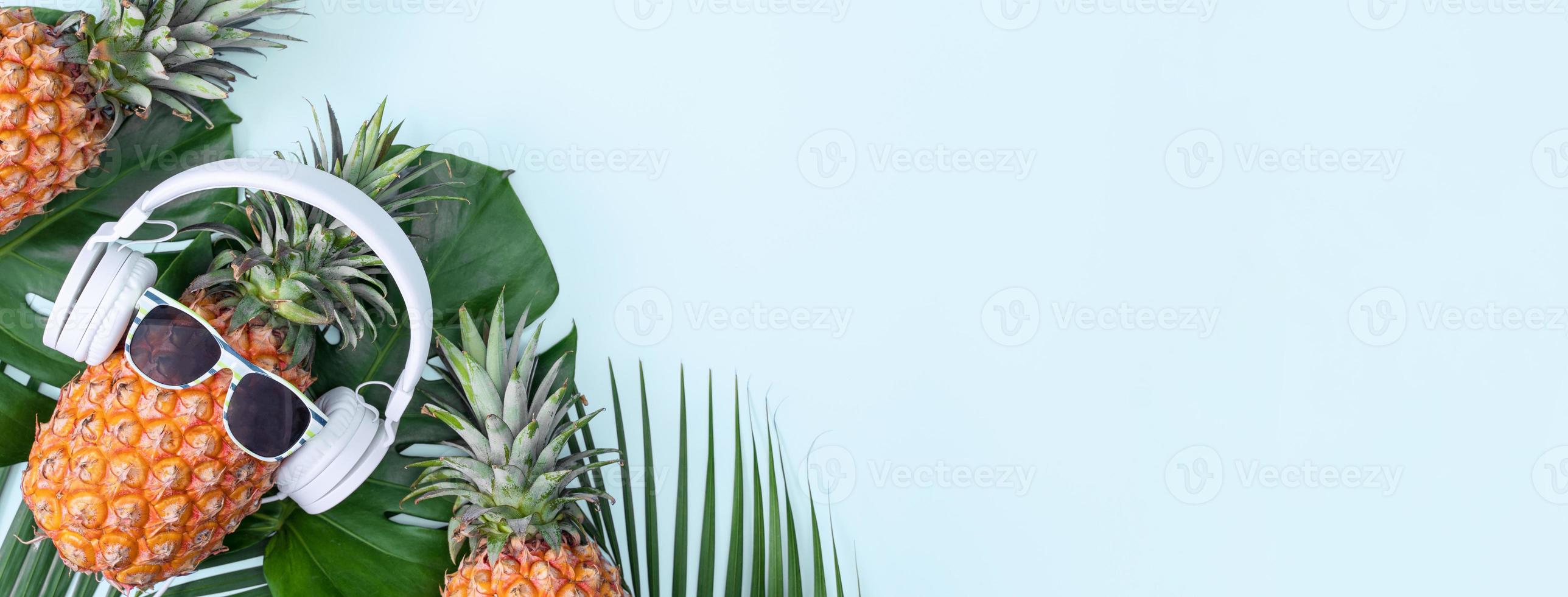 Funny pineapple wearing white headphone, concept of listening music, isolated on blue background with tropical palm leaves, top view, flat lay design. photo