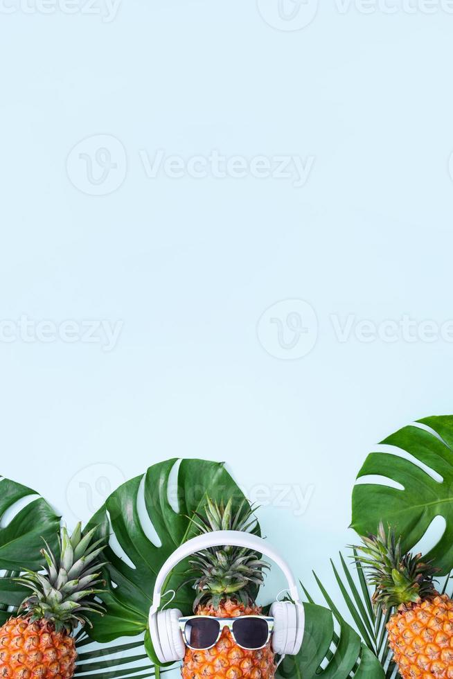 Funny pineapple wearing white headphone, concept of listening music, isolated on blue background with tropical palm leaves, top view, flat lay design. photo