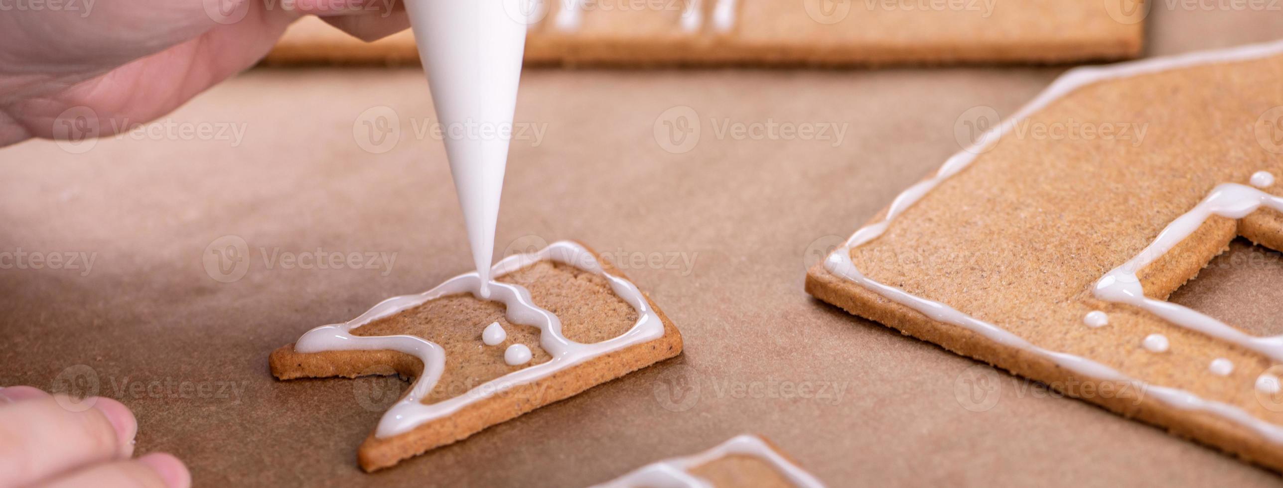 Young woman is decorating Christmas Gingerbread House cookies biscuit at home with frosting topping in icing bag, close up, lifestyle. photo